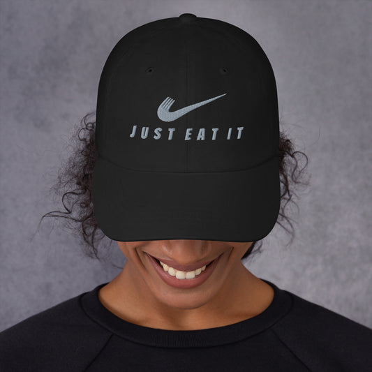 Black dad hat featuring a parody Nike logo and "Just Eat It" text, from Appetite Apparel with a classic curved brim.